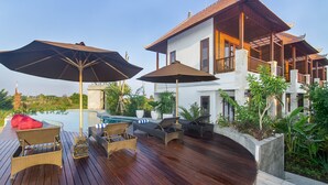 Terraza de madera con sillas de descanso y sombrillas, una piscina y una casa moderna con techo de tejas.