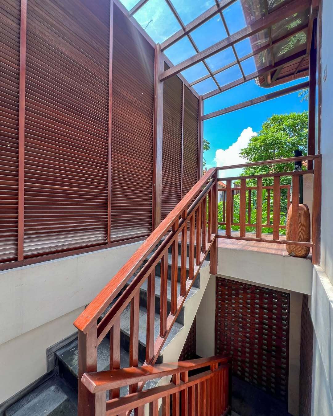 A staircase with wooden railings leading to a balcony with a glass ceiling, offering a view of greenery.