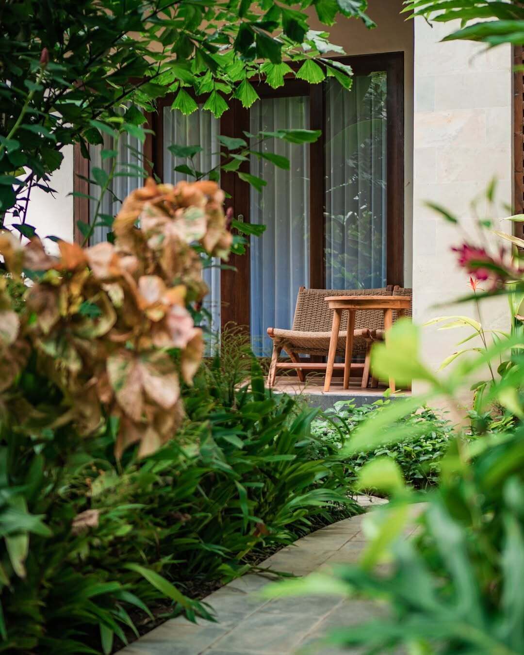 Un jardín con sillón de mimbre y mesa, rodeado de abundante vegetación y un sendero de piedra.