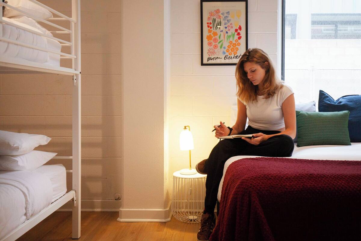 A room with a bunk bed, a single bed with a red blanket, a small table with a lamp, and a framed artwork on the wall.
