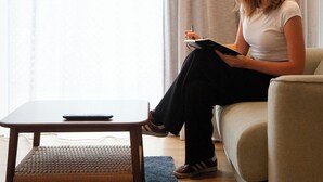 Una persona asseguda en un sofà amb un quadern i una ploma, una taula petita amb un teclat, i una finestra amb cortines.