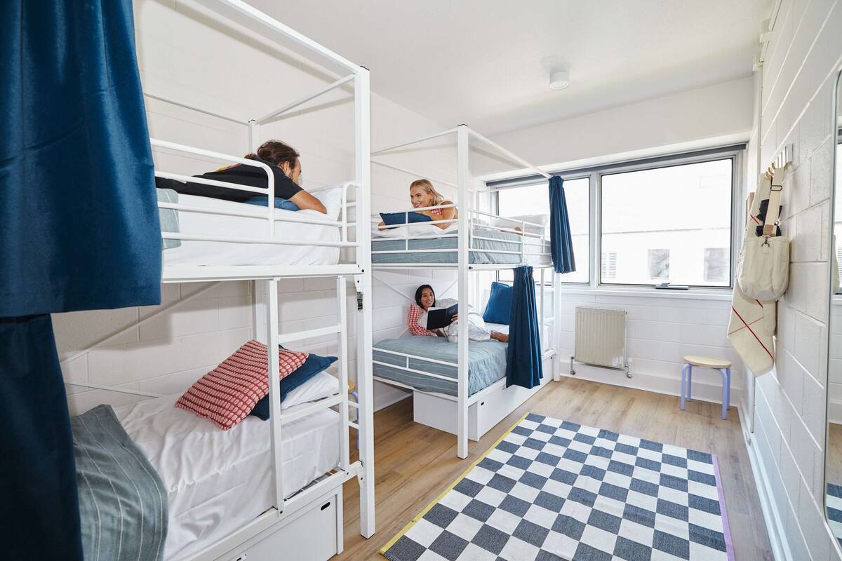 A dormitory room with bunk beds, a window with curtains, a radiator, and a checkered rug.