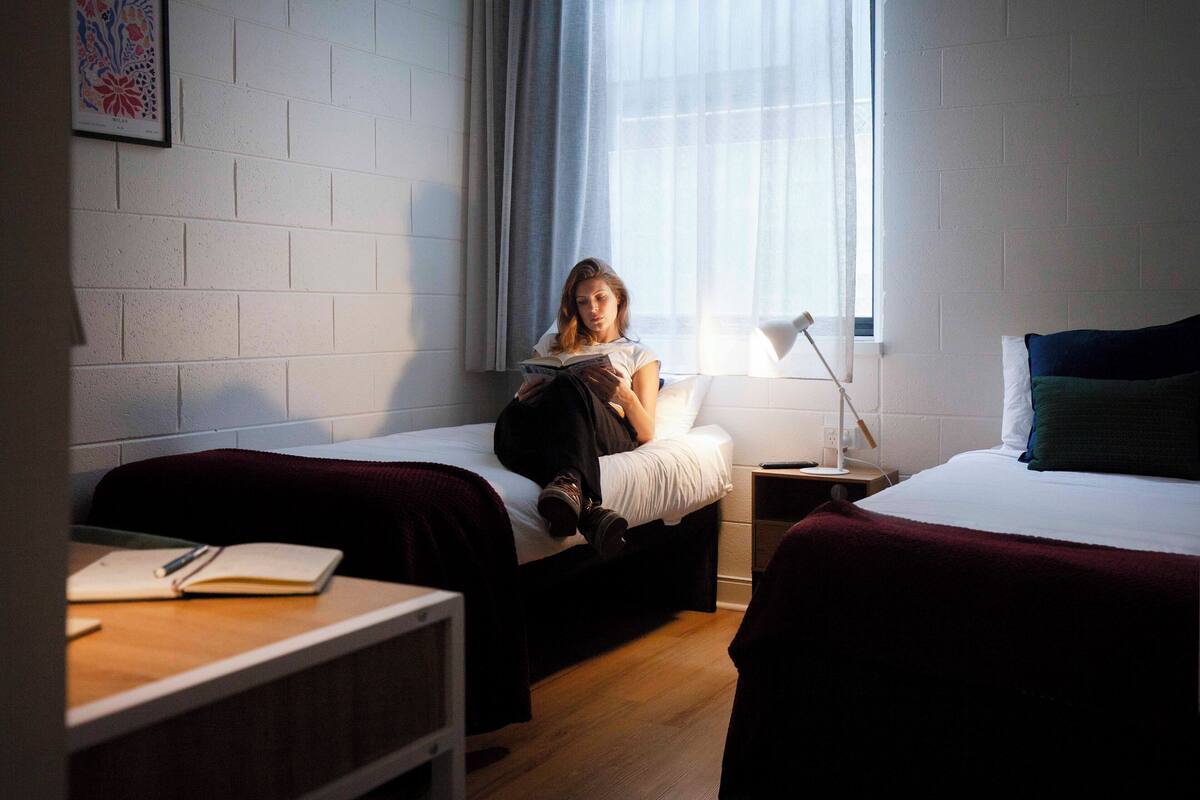 A room with two beds, a bedside table with a lamp, and a window with curtains.
