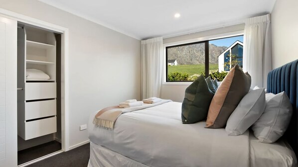Un dormitorio con cama, ventana con vista a una casa y follaje, y armario con estantes y cajones.