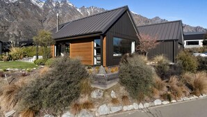 Una casa moderna con techo oscuro, revestimiento de madera y ventanas amplias. Está rodeada por un jardín ajardinado con arbustos variados y un borde de piedra.