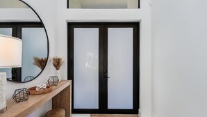 A modern entryway with a wooden console table, a round mirror, and a black-framed door with frosted glass panels. - Heated Saltwater I Pool Game Room | Firepit I BBQ (Cape Coral)