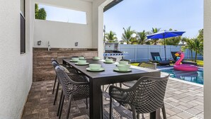 An outdoor patio with a dining table set for six, a grill, and a pool with a flamingo float. - Heated Saltwater I Pool Game Room | Firepit I BBQ (Cape Coral)