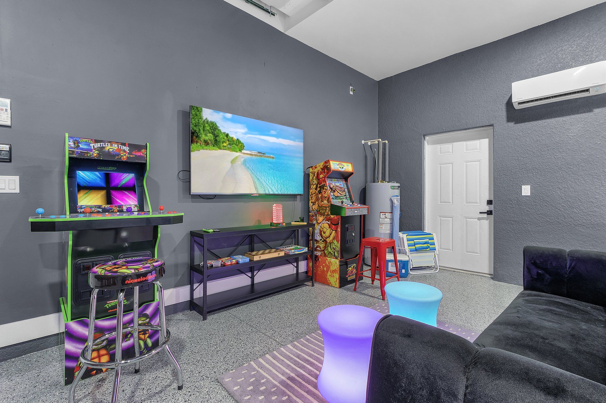 A room with a large flat-screen TV, a Nintendo Switch console, a colorful arcade machine, and a comfortable seating area with a couch and ottoman.