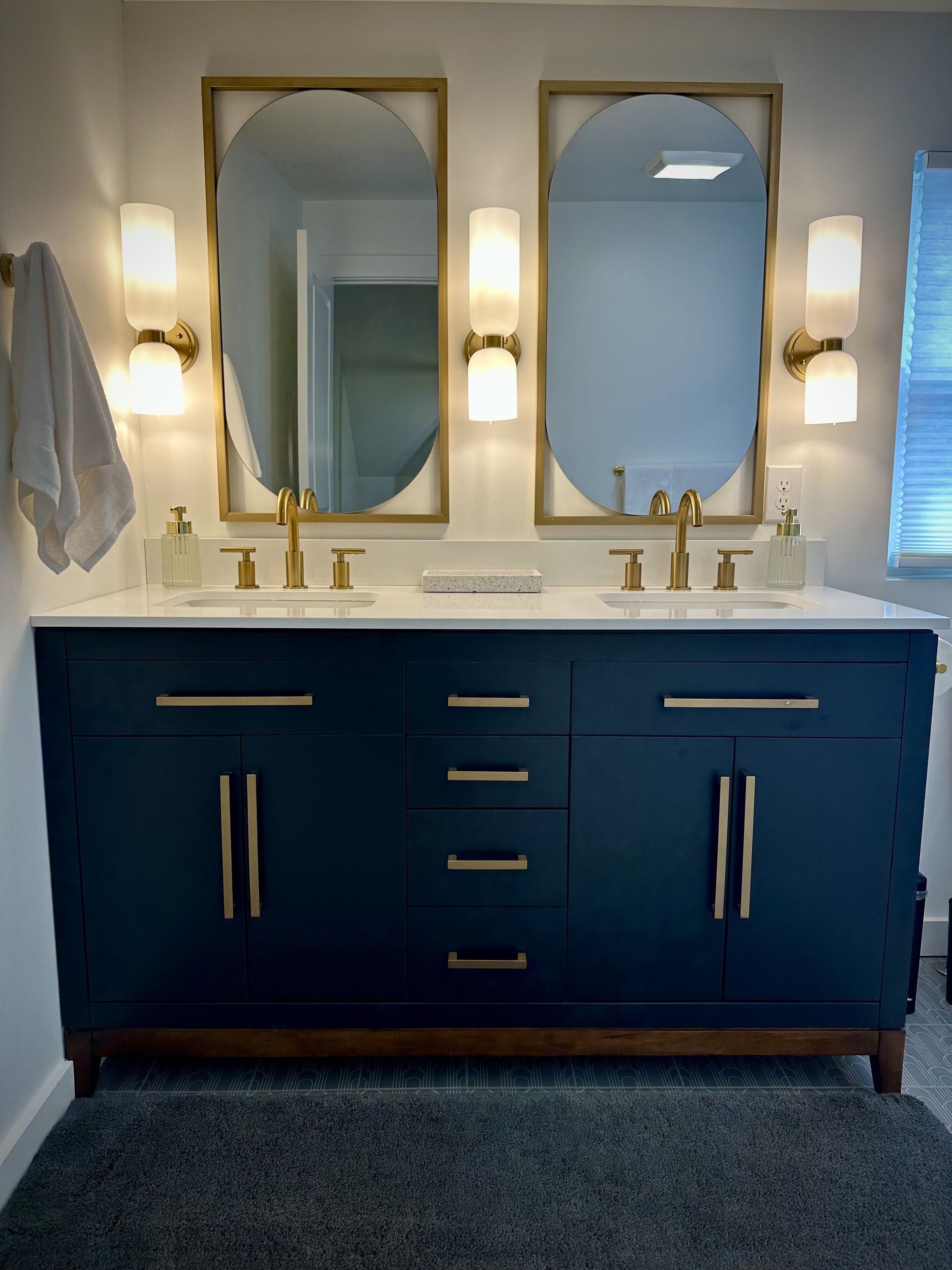 A bathroom with two sinks, gold faucets, and a dark blue vanity with gold handles. There are wall-mounted lights above each mirror.