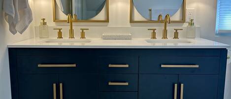 A bathroom with two sinks, gold faucets, and a dark blue vanity with gold handles. There are wall-mounted lights above each mirror.