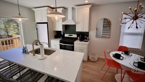 Une cuisine moderne avec des meubles blancs, un îlot central, un coin repas avec des chaises rouges et une grande fenêtre équipée de stores.