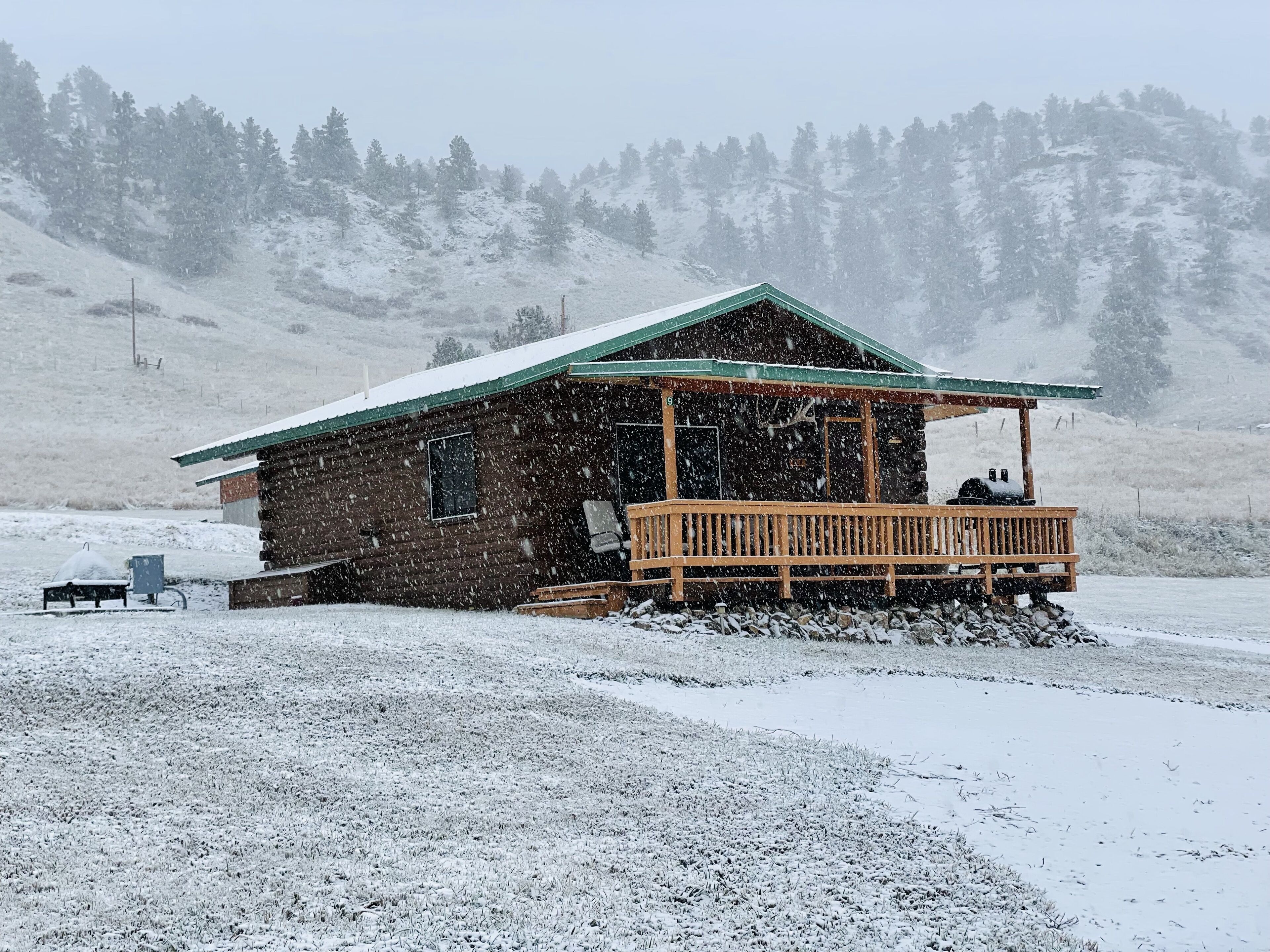 A wooden cabin with a porch, surrounded by a snowy landscape and a forested hill in the background.
