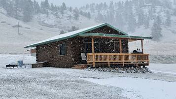 A wooden cabin with a porch, surrounded by a snowy landscape and a forested hill in the background.