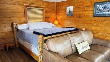 A wooden cabin room with a bed, a leather sofa, a lamp, and two framed pictures on the wall.