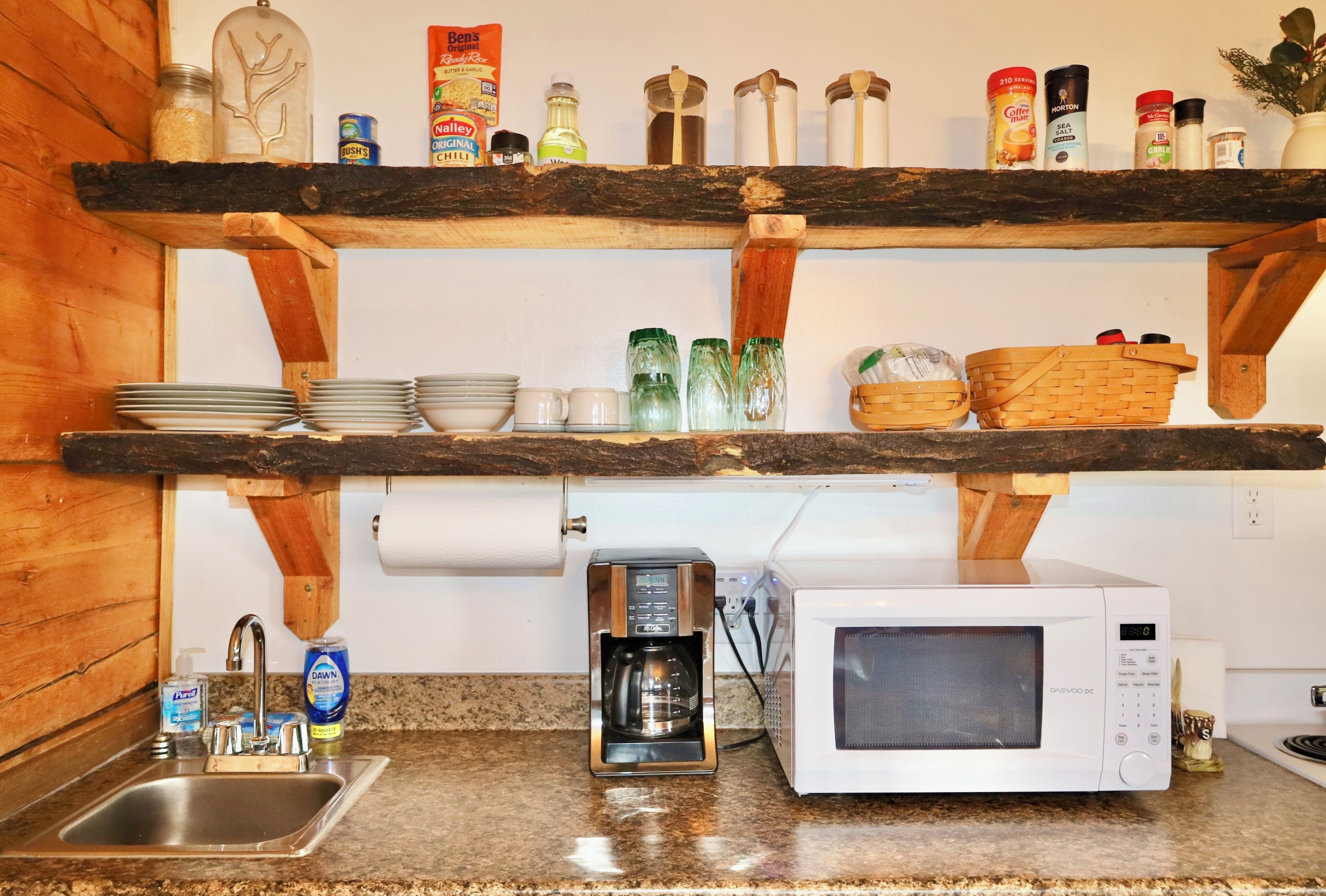 Une cuisine avec une étagère en bois où sont rangés divers ustensiles, une machine à café, un four à micro-ondes et un évier avec du liquide vaisselle.
