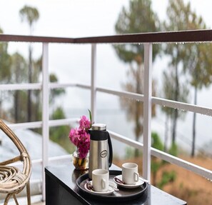 A balcony with a view of trees and water, a wicker chair, a tray with a coffee maker, cups, and a vase with flowers.