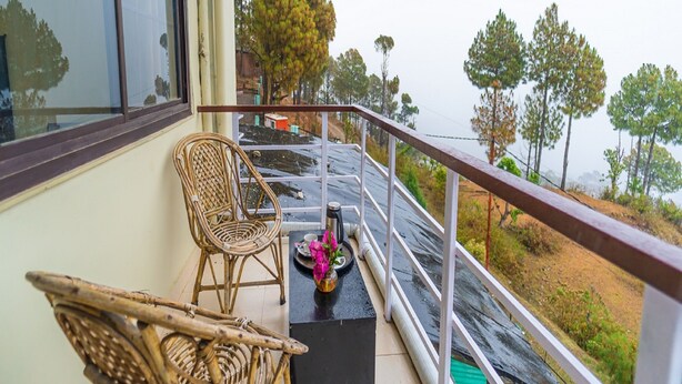 A balcony with wicker chairs, a small table with a vase, and a view of trees and a road. - Flutter Hotels & Resort (Lansdowne)