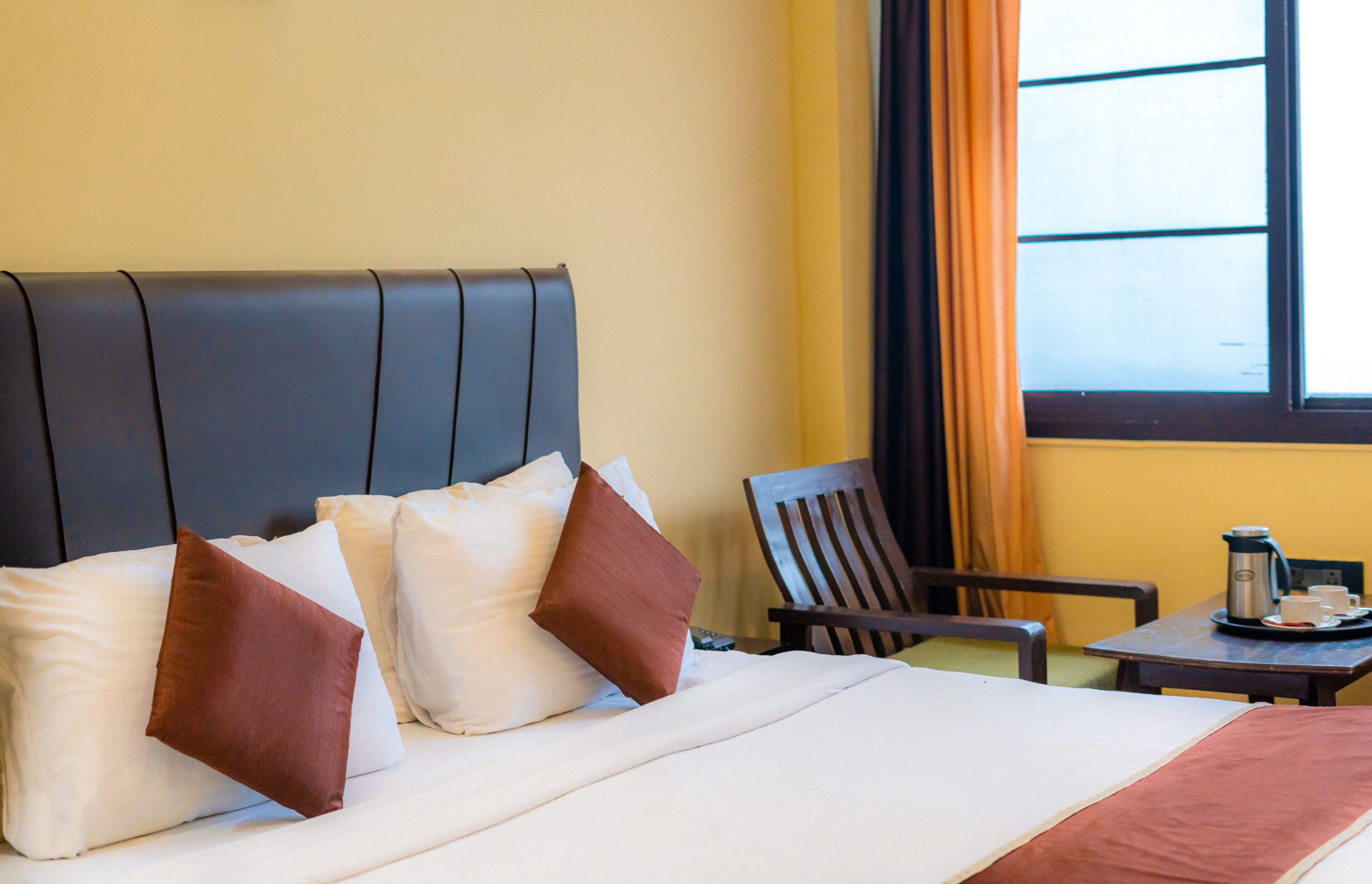 A hotel room with a bed, pillows, a chair, a table with a coffee pot, and a window with curtains.