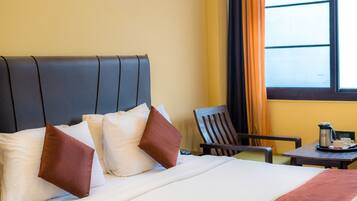 A hotel room with a bed, pillows, a chair, a table with a coffee pot, and a window with curtains.