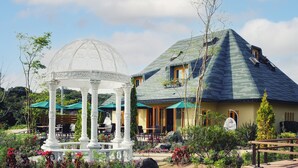 Un kiosque surmonté d’un dôme blanc et orné de colonnes, entouré d’un jardin où poussent diverses plantes et fleurs.