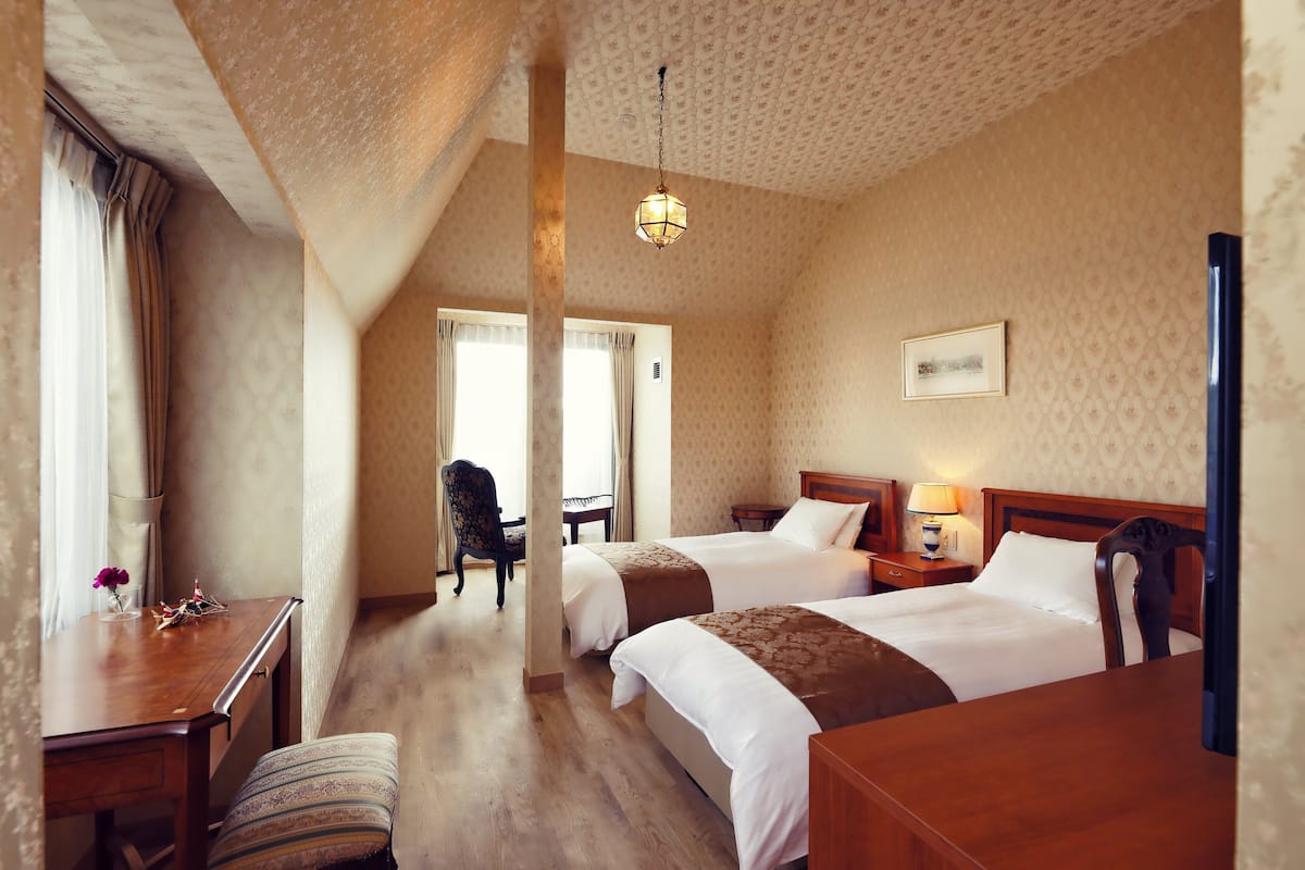 A hotel room with two beds, a desk, a chair, and a lamp.