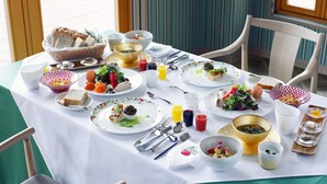 Une table à manger est dressée avec différents plats, couverts et verres, ce qui indique qu’un repas va avoir lieu dans une pièce dont les murs sont recouverts d’un papier peint à rayures et qui possède une fenêtre.