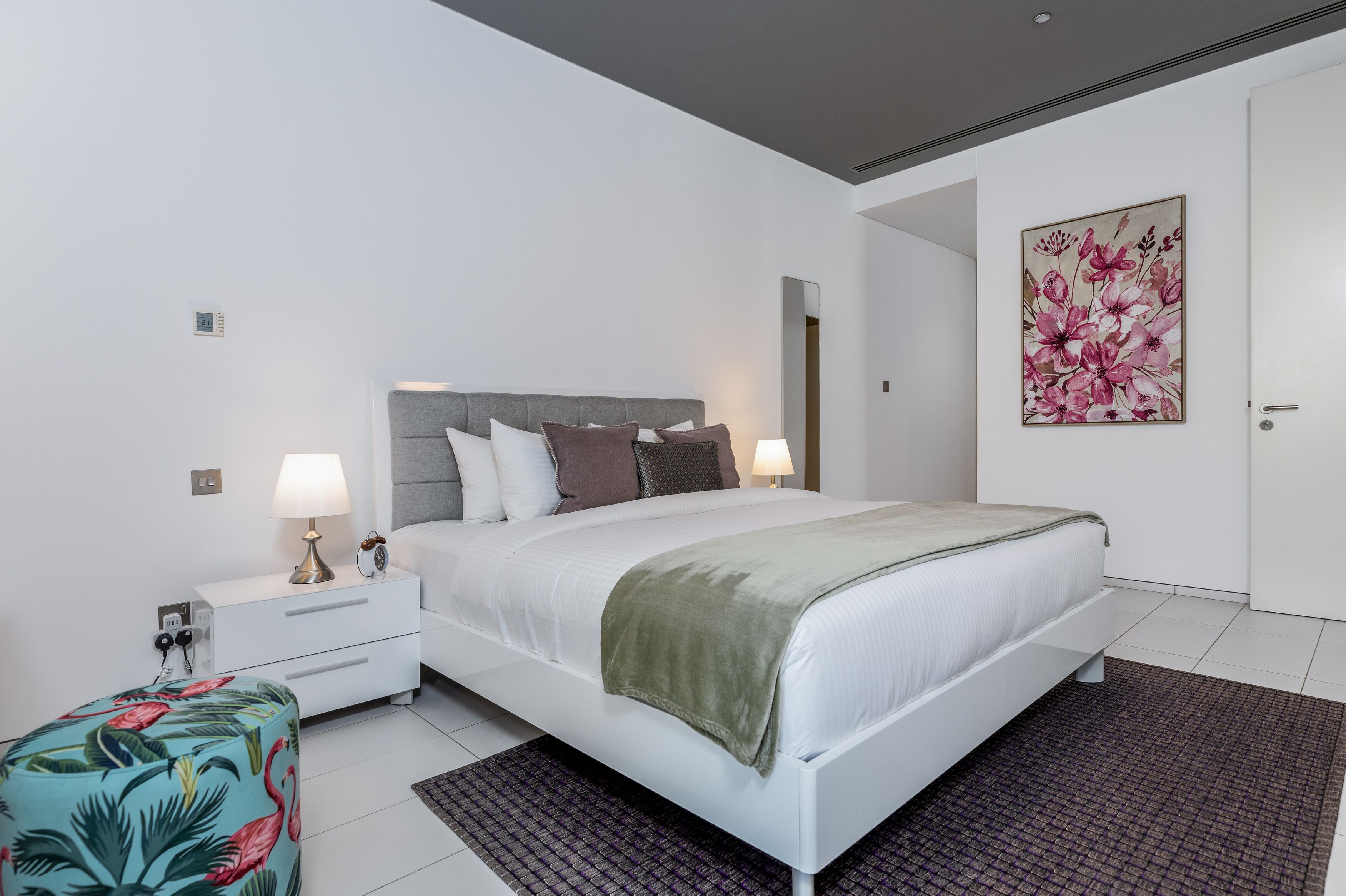 A modern bedroom with a white bed, grey headboard, and a floral painting on the wall.