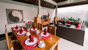 A dining area with a wooden table set for six, red napkins, and a vase of flowers. There is a sofa with red cushions in the background. - Taman Bali Villa Dewi Sri (Legian)