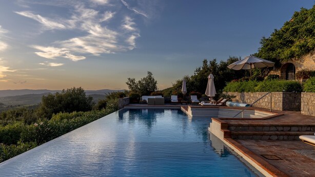 Ein Infinity-Pool mit einer Steindeck und Liegestühlen, umgeben von viel Grün und Blick auf ferne Hügel.