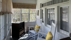 Una habitación acogedora con techo de madera, ventanas amplias con persianas, un banco de madera con cojines y una mesita con lámpara.