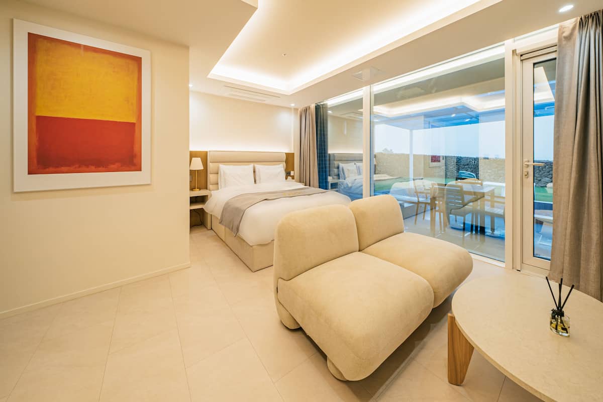A modern hotel room with a bed, a sofa, a table, and a balcony with outdoor seating.