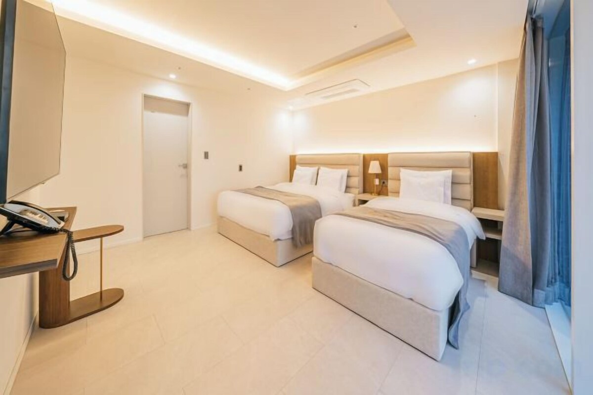 A hotel room with two beds, a desk with a telephone, and a large window with curtains.