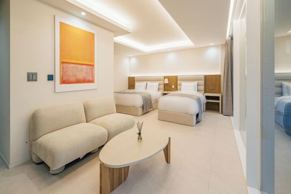 A modern hotel room with a sofa, two beds, a small table, and a painting on the wall.