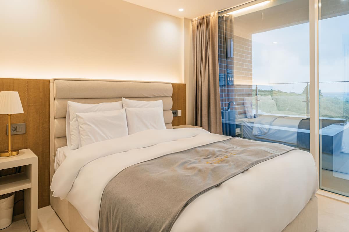 A modern hotel room with a large bed, a nightstand, and a view of the outdoors through a glass door.