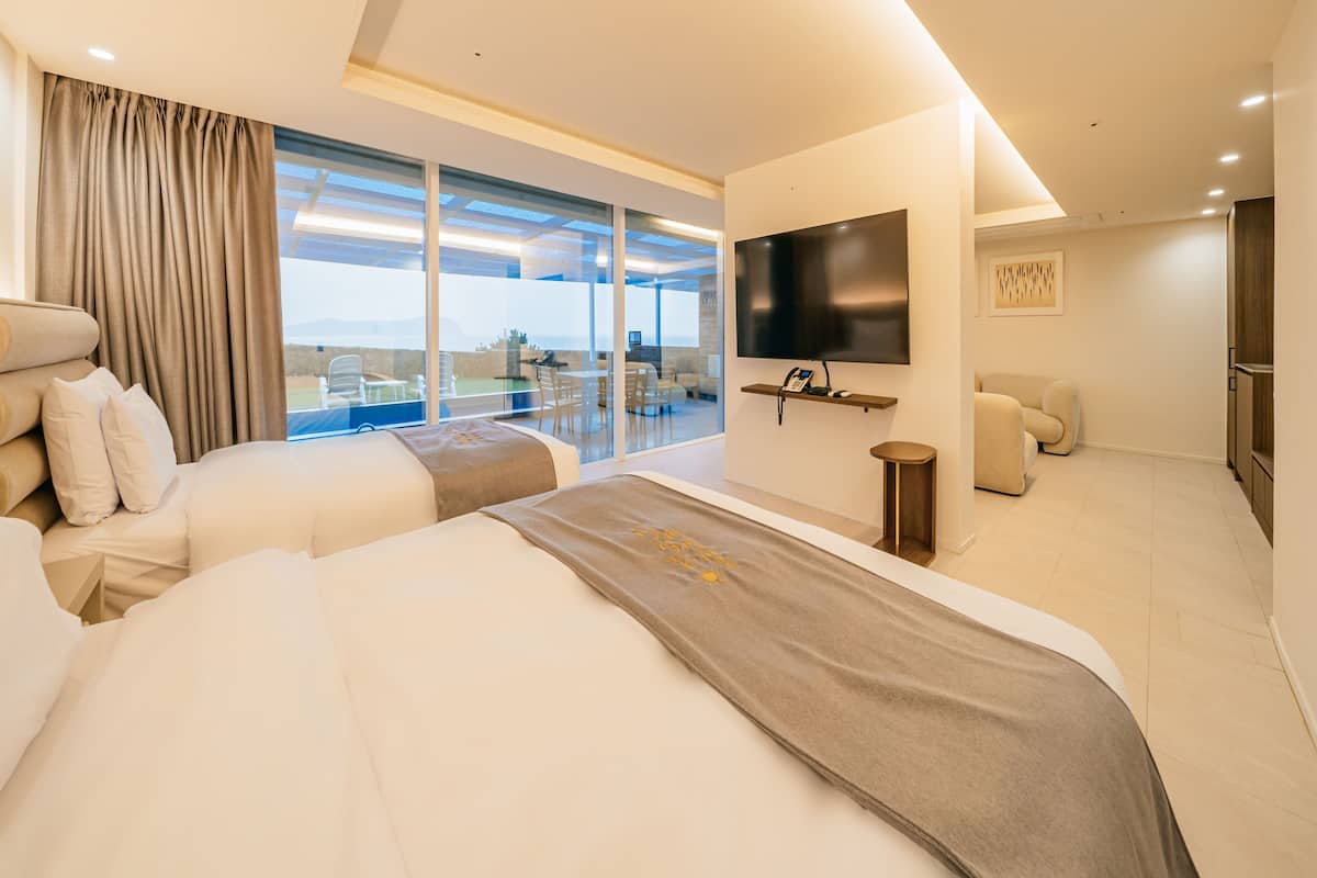 A modern hotel room with two beds, a TV, a balcony with a dining area, and a view of the sea.