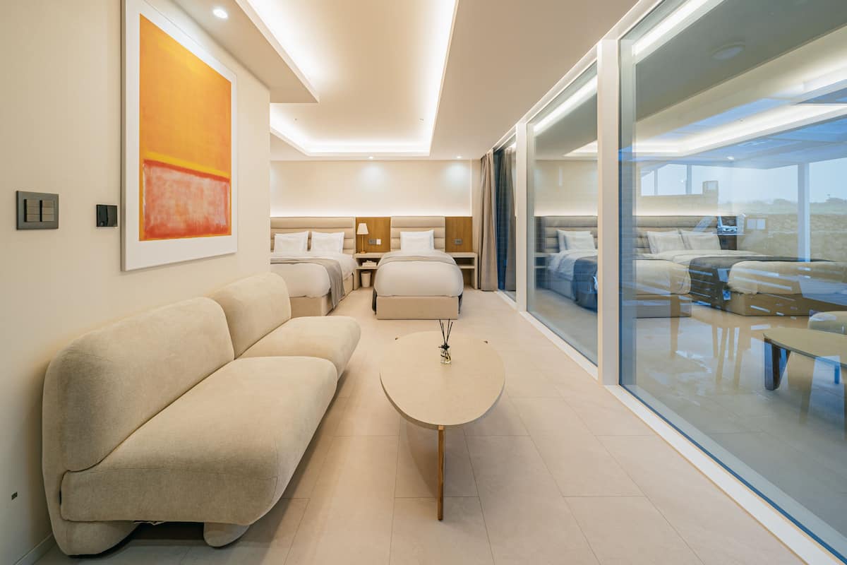A modern hotel room with a large bed, a sofa, a small table, and a large window offering a view of the desert.