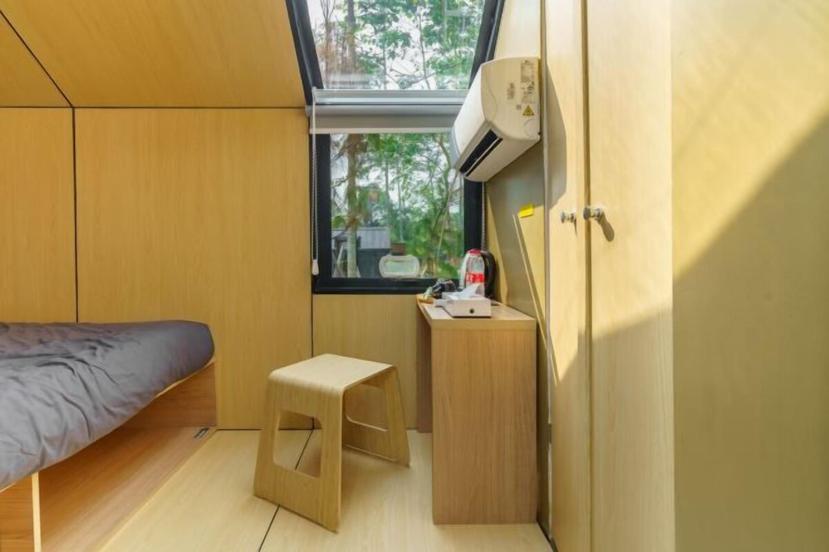 Una habitación pequeña con cama, un taburete, un escritorio y una ventana con aire acondicionado.