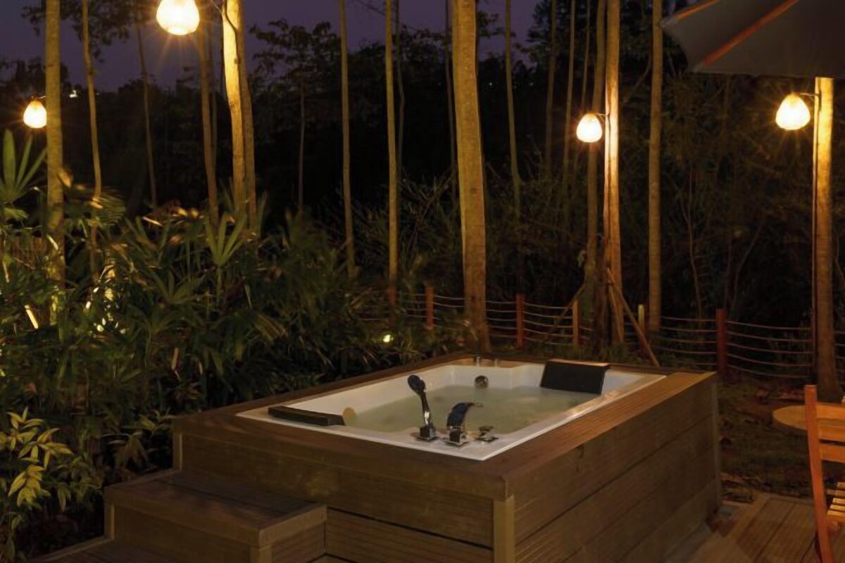 Tinaja de madera con asiento incorporado y una pequeña mesa, rodeada de vegetación exuberante y árboles altos.