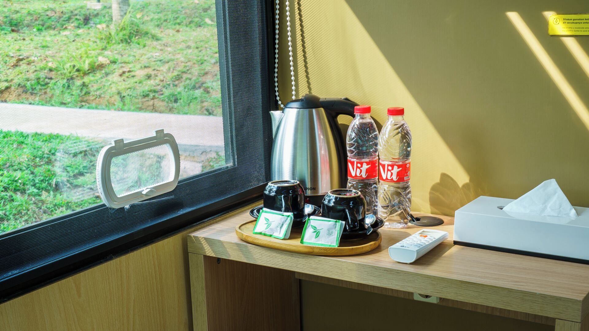 Een raam met uitzicht op groen, een waterkoker, twee kopjes, twee waterflessen, een zakdoos en een afstandsbediening op een houten oppervlak.