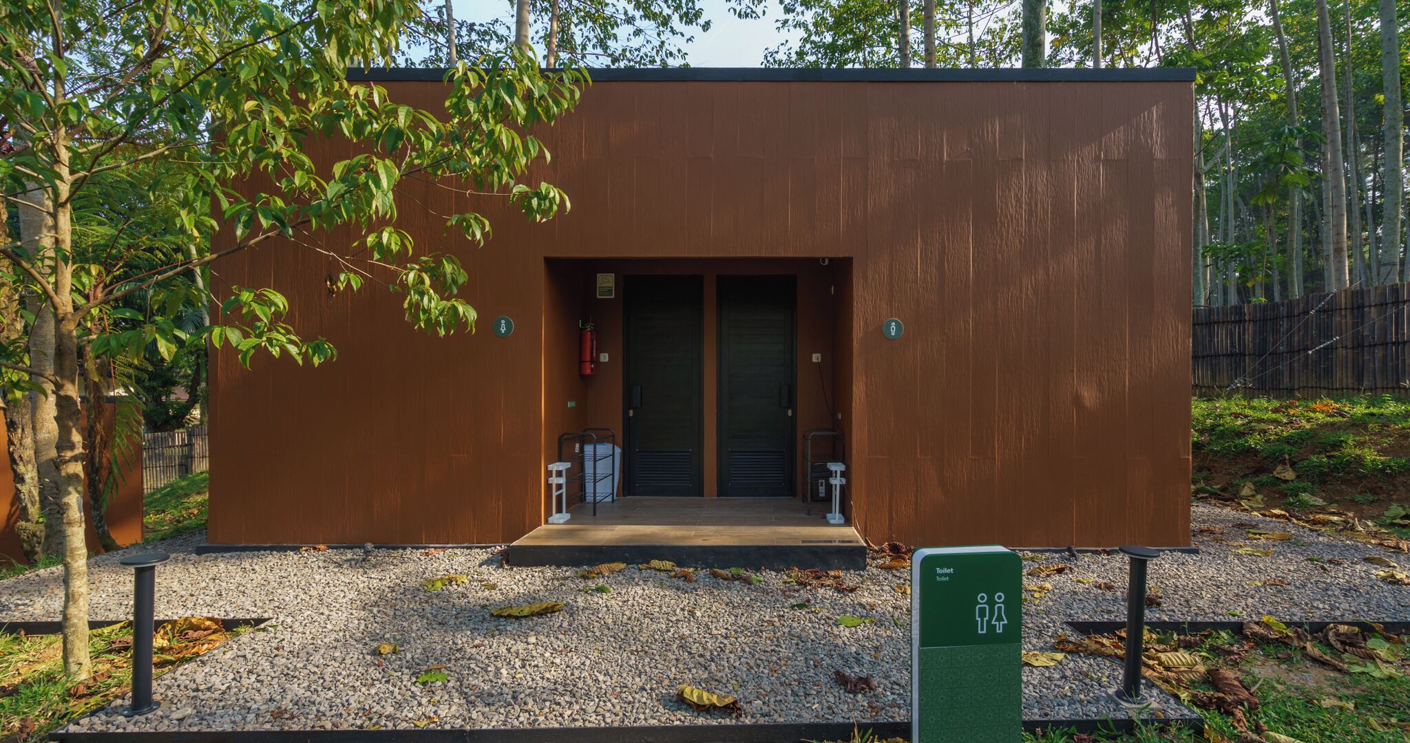 A modern, single-story building with a brown exterior, a door, and a small entrance area with a bench and a trash bin.