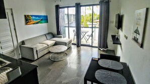 Ein modernes Wohnzimmer mit Sofa, Couchtisch und Esstisch. Es gibt einen Deckenventilator, ein Bild an der Wand und einen Ausblick ins Freie durch große Fenster.