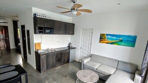 Eine moderne Küche mit dunklen Schränken, einer Mikrowelle und einer Kaffeemaschine. Ein graues Sofa und ein runder Tisch mit Drahtgestell sind ebenfalls vorhanden. Über der Küche ist ein Deckenventilator zu sehen.