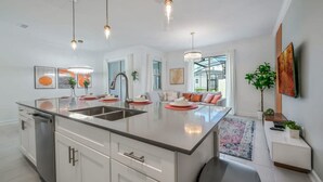 Une cuisine moderne dotée d’un îlot central, d’appareils électroménagers en acier inoxydable, de luminaires suspendus et offrant une vue sur l’extérieur.