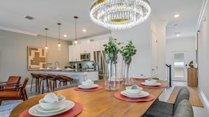 Un espace de repas moderne avec une table en bois dressée pour quatre personnes, entourée de chaises. Un lustre est suspendu au-dessus, et la pièce présente un design épuré et minimaliste, avec des murs blancs et une cuisine en arrière-plan.