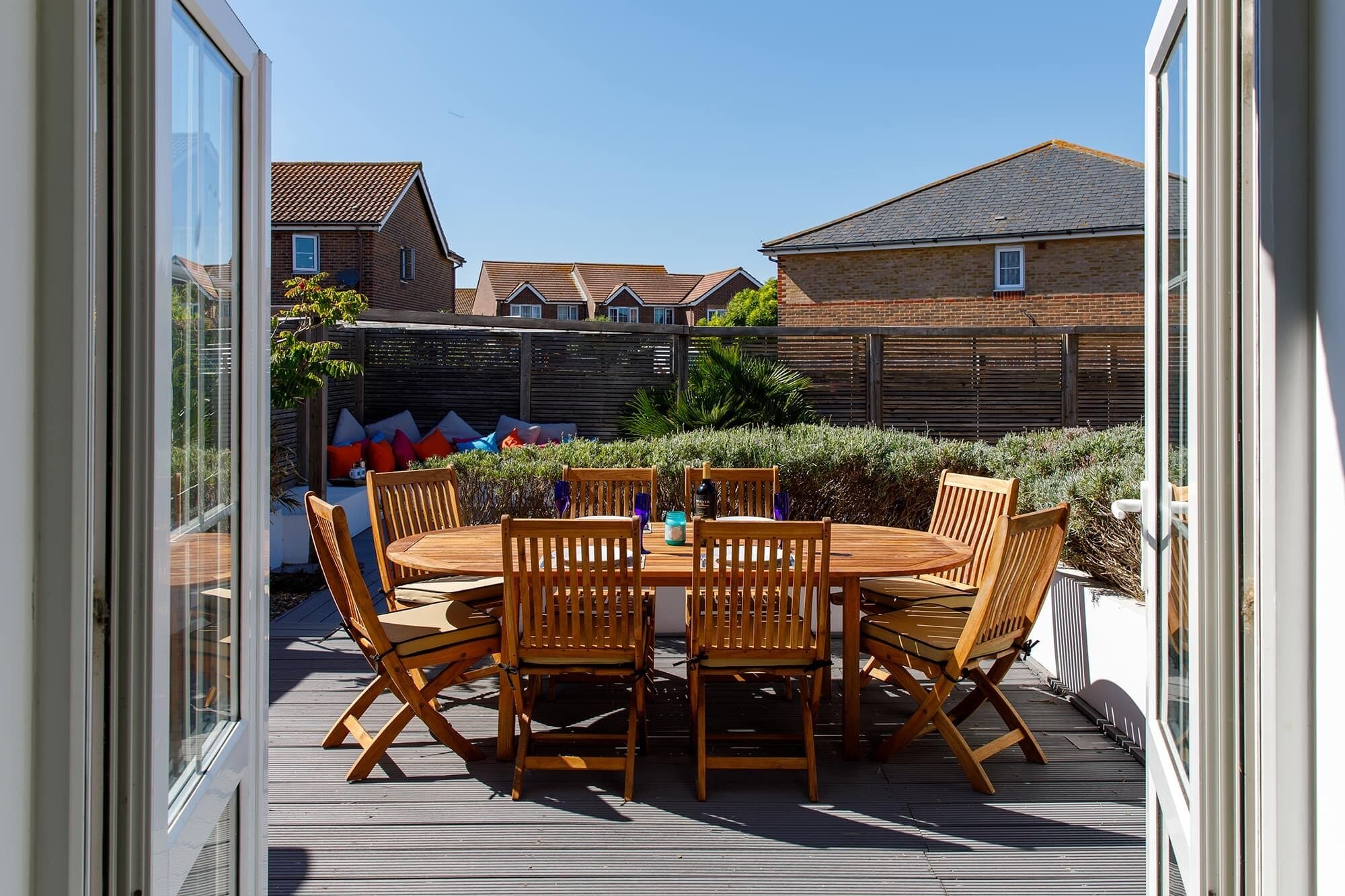 An outdoor patio with a wooden table and chairs, surrounded by a wooden deck and a low brick wall.
