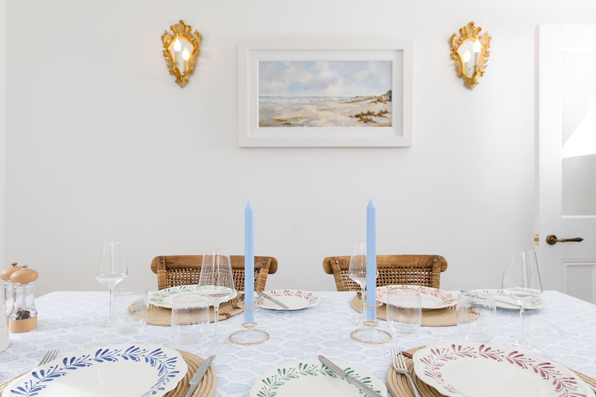 A dining table set with plates, glasses, and candles, flanked by wicker chairs and a framed painting on the wall.