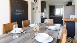A dining area with a wooden table set for four, a chalkboard with a welcome message, and a modern kitchen in the background. - Coastal Retreat for 6 Close to Beaches (East Wittering)