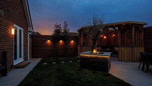 A backyard with a stone fire pit, wooden fence, and a wooden pergola with lights. - Coastal Retreat for 6 Close to Beaches (East Wittering)