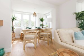 Quarto claro e arejado com área de jantar, destacando cadeiras de vime e uma mesa redonda, um sofá bege e grandes janelas que proporcionam iluminação natural.