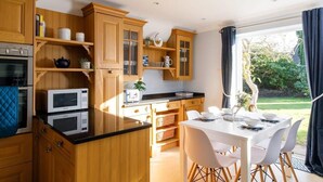 A modern kitchen with wooden cabinets, a dining area with a white table and chairs, and a view of a garden through a large window.